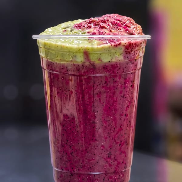Vibrant pink and green layered smoothie in a clear plastic cup on a dark surface.